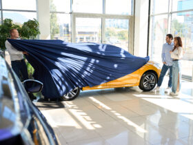 car salesman pulling off car cover to reveal car to young happy couple