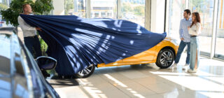 car salesman pulling off car cover to reveal car to young happy couple