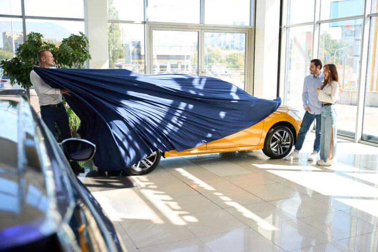 car salesman pulling off car cover to reveal car to young happy couple