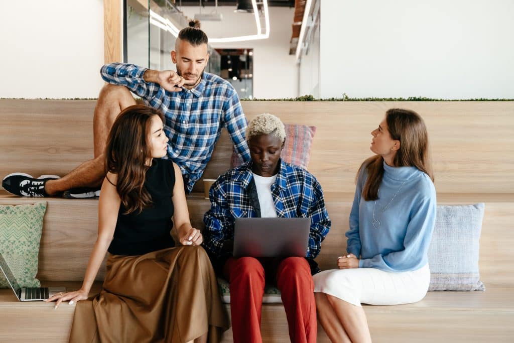 four people sitting on a bench, working together on a laptop