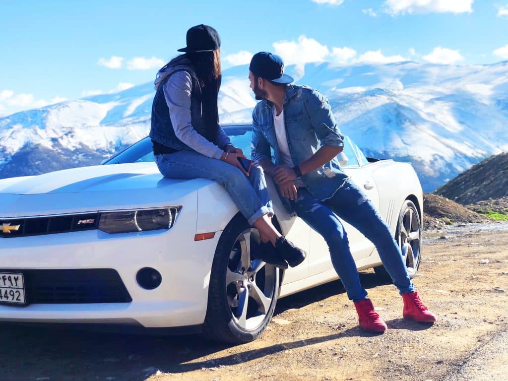 man and woman leaning and sitting on a white Chevy sports car that is parked on a hill overlooking a mountain range.