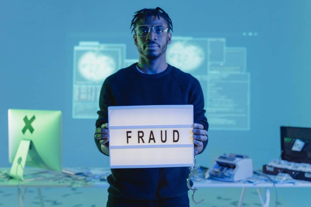 a man standing in front of a cluttered desk, holding a sign that says "Fraud"