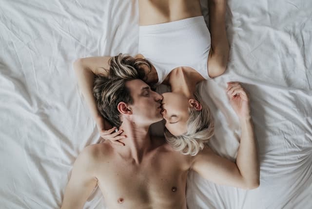 Couple laying in opposite directions on a bed with white sheets, kissing.