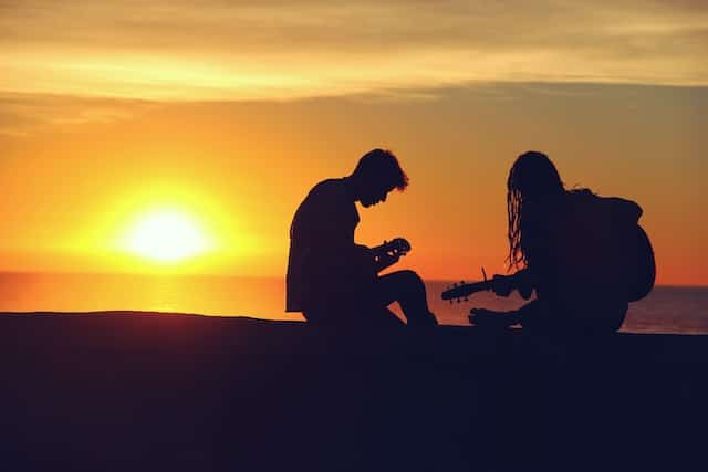 a couple both playing guitar while sitting on the beach at sunset