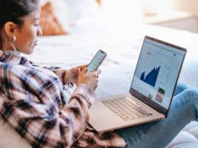 woman sitting with laptop in her lap and phone in her hands, looking at investment data and charts