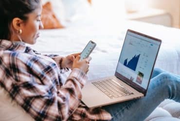 woman sitting with laptop in her lap and phone in her hands, looking at investment data and charts