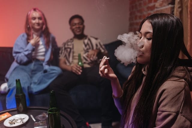 woman in brown jacket smoking a joint while at a party with two other people in the background.