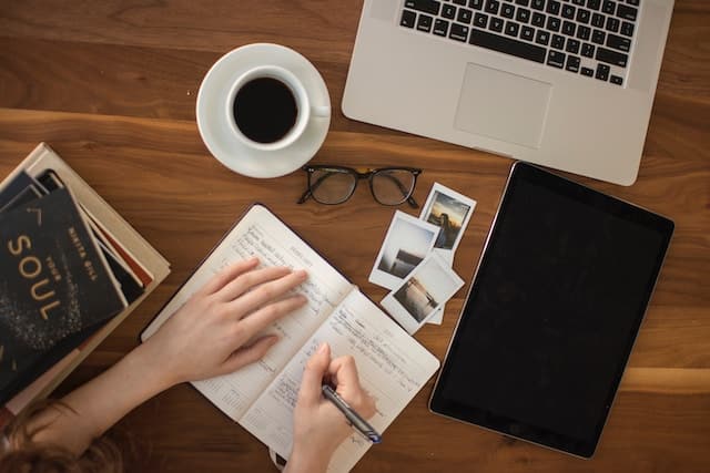 woman writing in a notebook with a cup of coffee and a laptop on her desk