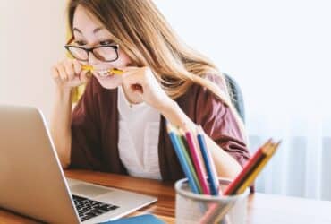 woman looking at a laptop in excitement and anticipation, while chewing on a pencil