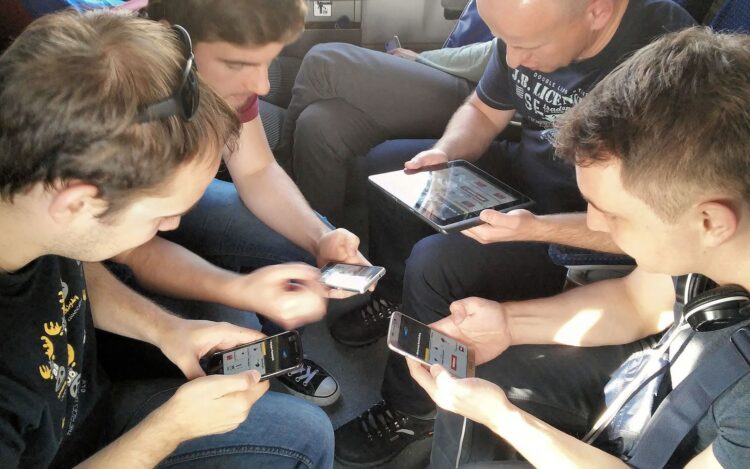 a group of guys sitting and playing a game together on their devices. Source: https://commons.wikimedia.org/wiki/File:Spaceteam_multiplayer_mobile_game.jpg