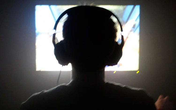 young man sitting in a dark room, playing a video game while wearing headphones
