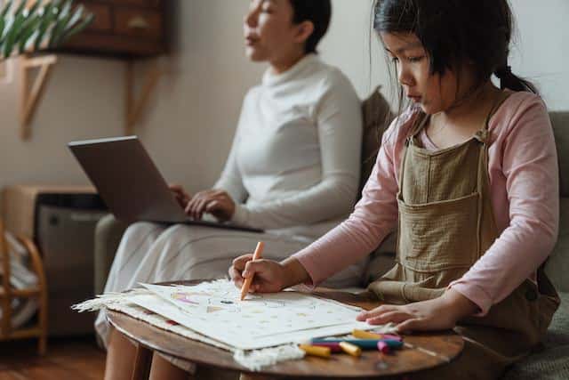 a young girl focused on coloring while her mother is working in the background