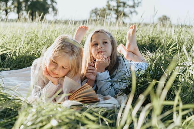 two girls laying in the grass and reading a book together