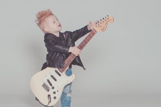 a young boy in a leather jacket holding an electric guitar that is too big for him