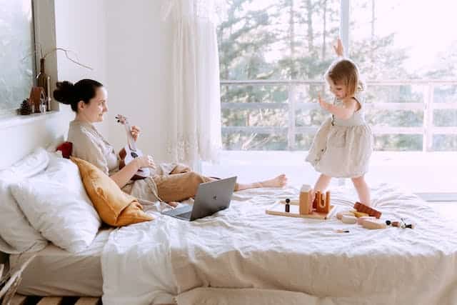 a mother playing a musical instrument on the bed while her young daughter dances on the bed