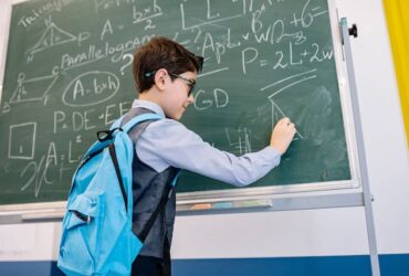 Boy wearing backpack working complex math problems on a chalkboard.