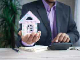 man holding a small model house in one hand while working on a calculator in the other.