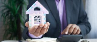 man holding a small model house in one hand while working on a calculator in the other.