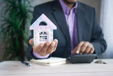 man holding a small model house in one hand while working on a calculator in the other.